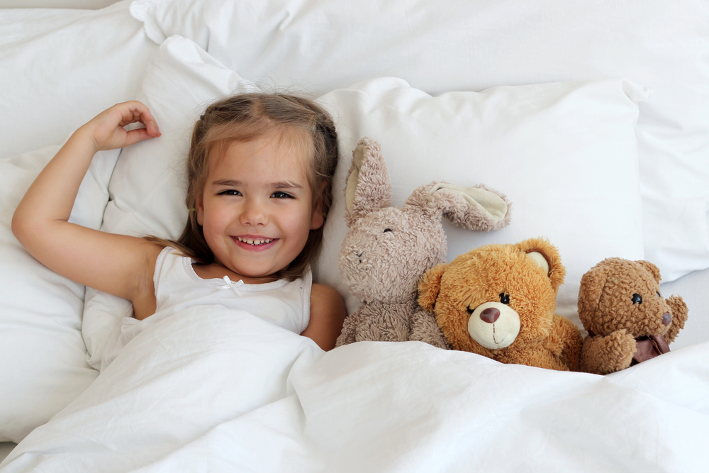 Dievča s plyšovými hračkami ležiace v povlečenej posteli s chráničom matraca